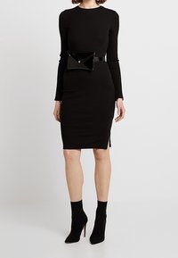Schwarzes geripptes Kleid mit langen Ärmeln, knielangem Schnitt, seitlichem Schlitz und einer glänzenden schwarzen Lackgürteltasche mit spitzem Klappen-Detail.