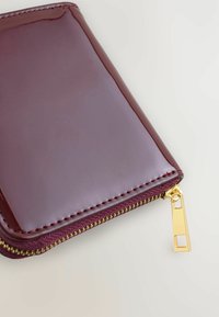 Portafoglio bordeaux lucido con zip dorata e cuciture bordeaux, caratterizzato da una texture liscia e forma rettangolare. Il tirazip è rettangolare.