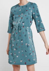 Tealfärgad klänning med rund halsringning, 3/4-långa ärmar och midjeband. Har ett blommönster i rött, rosa och mörkgrönt. Slät textur.