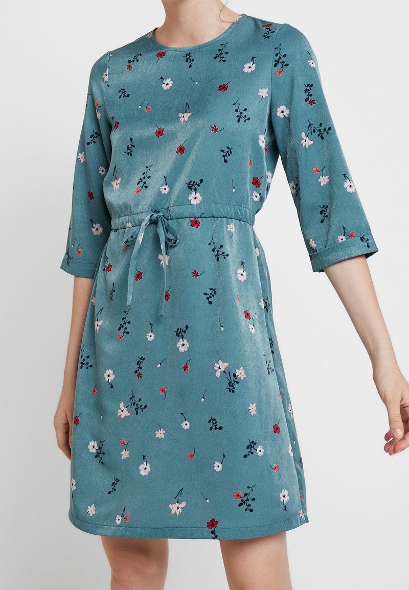Tealfärgad klänning med rund halsringning, 3/4-långa ärmar och midjeband. Har ett blommönster i rött, rosa och mörkgrönt. Slät textur.