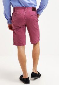 Men's shorts in berry pink, made of a lightweight fabric. Features two back pockets and a belt loop, paired with black slip-on shoes.