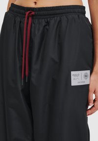 Czarne spodnie typu windbreaker z elastycznym paskiem w kolorze czerwonym, bocznymi kieszeniami oraz białą metką z napisem na lewym udzie.