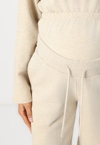 Pantalons de maternité beige clair en matière molletonnée, avec ceinture élastique, détail de cordon de serrage et texture douce, associés à un haut assorti à manches longues.