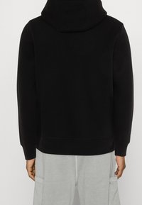 Sudadera negra con bolsillo tipo canguro, puños y dobladillo acanalados. Hecha de material suave, cuenta con capucha y un corte relajado. Lleva puesta sobre pantalones de chándal grises.