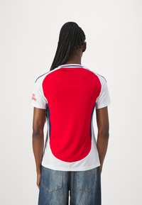Červený a bílý atletický dres s modrými akcenty. Má krátké rukávy, kruhový výstřih a strukturovanou tkaninu. Zadní pohled ukazuje logo na rukávu.