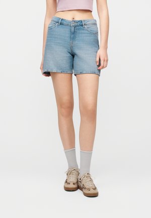 Donna che indossa pantaloncini di jeans azzurri, calzini grigi, sneakers beige e una maglietta corta rosa, in piedi davanti a uno sfondo bianco.