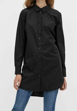 Kvinde iført en lang sort skjorte med knapper, krave og lange ærmer, kombineret med blå jeans, stående mod en ensfarvet baggrund.