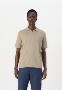 Camisa polo beige con cuello clásico, mangas cortas y una tela texturizada. Combinada con pantalones azules; diseño minimalista y colores neutros.
