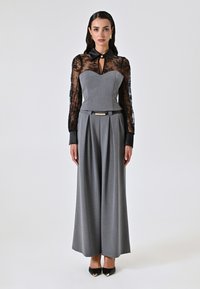 Grauer Jumpsuit mit strukturiertem Oberteil und langen, transparenten schwarzen Spitzenärmeln. Besitzt einen Kragen, einen Ausschnitt in Form eines Schlüssellochs und weit geschnittene, plissierte Hosenbeine.