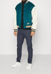 Teal- och krämfärgad collegejacka med en kropp av ull och ärmar av läder. Har randig ribbning och en liten logotypapplikation. Matchas med mörka byxor och vita sneakers.