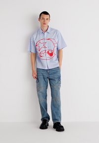 Chemise à manches courtes, bleu clair, à boutons avec un design graphique rouge ; associée à un jean cargo ample, bleu clair, et des chaussures noires imposantes.