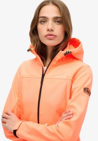 Veste softshell orange néon avec capuche, fermeture éclair sur toute la longueur et accents de logo. Présente une finition texturée et des poignets élastiques pour un ajustement optimal.