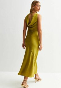 Klänning med känguru-hals i olivgrön färg, tillverkad av ett mjukt tyg och med en figurnära silhuett samt flytande nederkant. Har en öppen rygg med justerbara band.