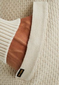 Beiger Pullover mit strukturiertem Strickmuster, featuring gerippte Bündchen und einem eingenähten Etikett. Eine Hand ruht in der Tasche, die Hautfarbe und -struktur zeigend.