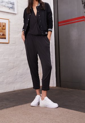 Woman wearing black jacket, black wrap top, black loose pants, and white sneakers standing indoors with hands in pockets.