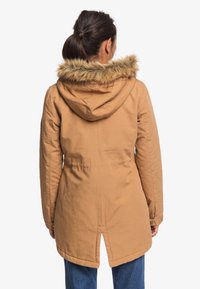 Giacca parka marrone con cappuccio in pelliccia sintetica. Presenta una vita aderente, maniche lunghe e una apertura sul retro per facilitare i movimenti. Texture liscia.