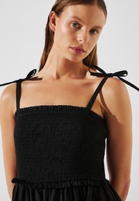 Il vestito nero presenta un corpino arricciato, spalline sottili con dettagli a fiocco e un orlo con volant, realizzato in tessuto leggero.