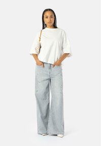T-shirt bianco oversize, jeans a gamba larga azzurri chiari con dettagli strappati, e una piccola borsa beige a scacchi. Outfit semplice e casual.