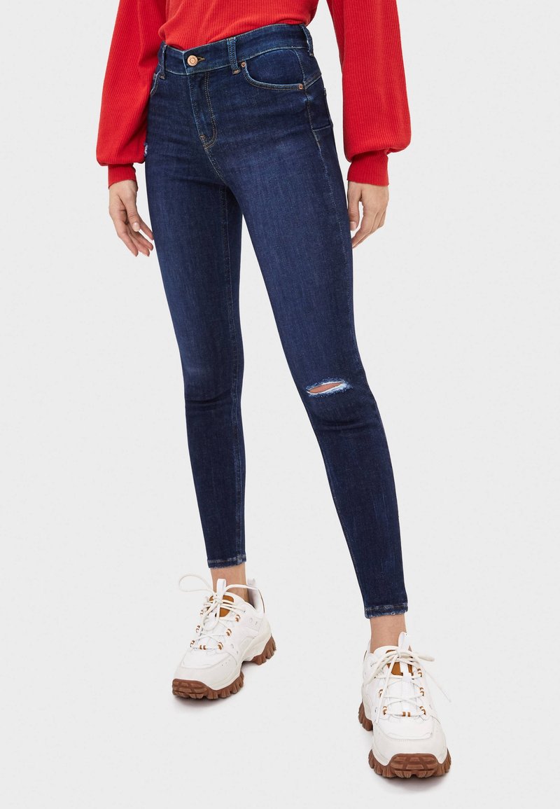 Jean skinny taille haute bleu foncé avec un genou déchiré. Assortis à des baskets blanches avec des semelles marron et des accents orange.