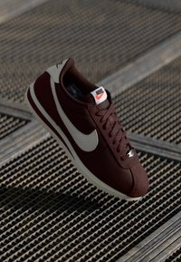 Scarpa da allenamento bordeaux con superficie texturizzata, baffo Nike bianco, lacci neri e suola bianca. Presenta un logo sulla linguetta.