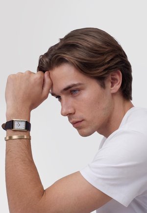 Nuori mies valkoisessa paidassa lepuuttaa päätään nyrkillä, ranteessa on suorakaiteen muotoinen kello mustalla hihnalla ja kultainen rannekoru.