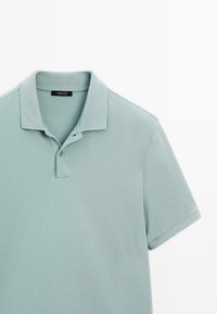 Ljus teal polo-tröja gjord av texturerad bomull. Har en klassisk krage, tvåknappslå och korta ärmar. Etikett synlig inuti kragen.