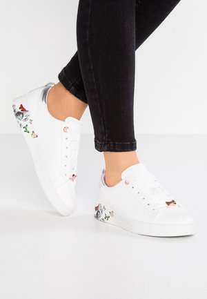 Personne portant des baskets blanches avec des motifs colorés de papillons et de fleurs sur le côté, associées à un jean skinny noir.
