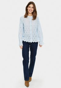 Blusa azzurra chiara con dettagli in pizzo, maniche lunghe e orlo a onde, abbinata a jeans blu scuro e stivali con tacco color cammello.