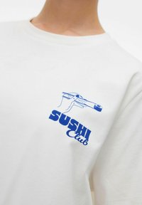 Biała koszula z niebieską grafiką ręki trzymającej pałeczki oraz napisem "SUSHI Club" na piersi.