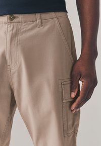Pantalon cargo beige en tissu robuste, avec une poche latérale, une taille ajustée et des détails de couture apparents. La main visible tient le tissu.
