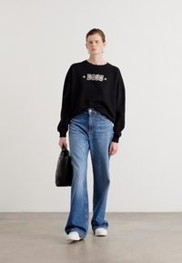 Felpa nera con logo "BOSS", jeans svasati blu, scarpe da ginnastica bianche, che tiene una borsa nera testurizzata. Sfondo semplice, modello in piedi dritto.