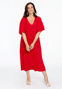 Rode, geplooide maxi-jurk met een diepe V-hals en vleermuismouwen, gecombineerd met witte strappy hakken. Soepele textuur, vloeiende uitstraling.
