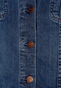 Jeansplagg med blå tvätt, som har fem bronsknappar med ingraverat varumärkesnamn, synliga ton-i-ton-sömmar och en distinkt zickzack-söm.