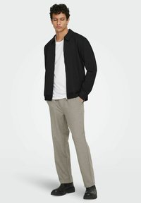 Mann som står med hendene i lommene, iført en svart jakke med glidelås, hvit t-skjorte, grå bukser og svarte støvler på en ensfarget bakgrunn.