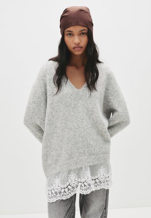 Un pull en peluche gris avec un col en V, porté sur un haut blanc transparent avec des finitions en dentelle. Le mannequin porte également un foulard marron.
