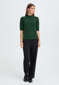 Pull en maille vert à manches courtes avec un col montant, associé à un pantalon large noir et des chaussures noires, sur un fond neutre.