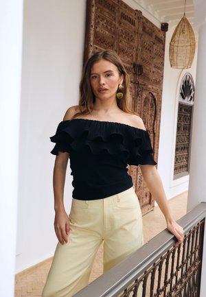 Jeune femme aux longs cheveux bruns portant un haut noir à épaules dénudées et volanté, et un pantalon jaune clair, debout près d'une porte en bois décorative.
