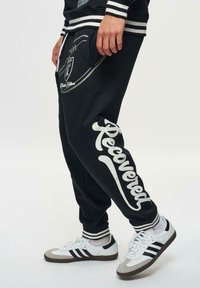 Zwarte trainingsbroek met witte graphics met de tekst "Hersteld." Beschikt over een voetballogo op de zak en gestreepte geribbelde manchetten. Gecombineerd met witte sneakers.
