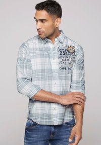 Hellblau-weiß kariertes Hemd mit Knopfkragen, hochgerollten Ärmeln und bedrucktem Textdetails auf der Vorderseite. Getragen mit Jeans.