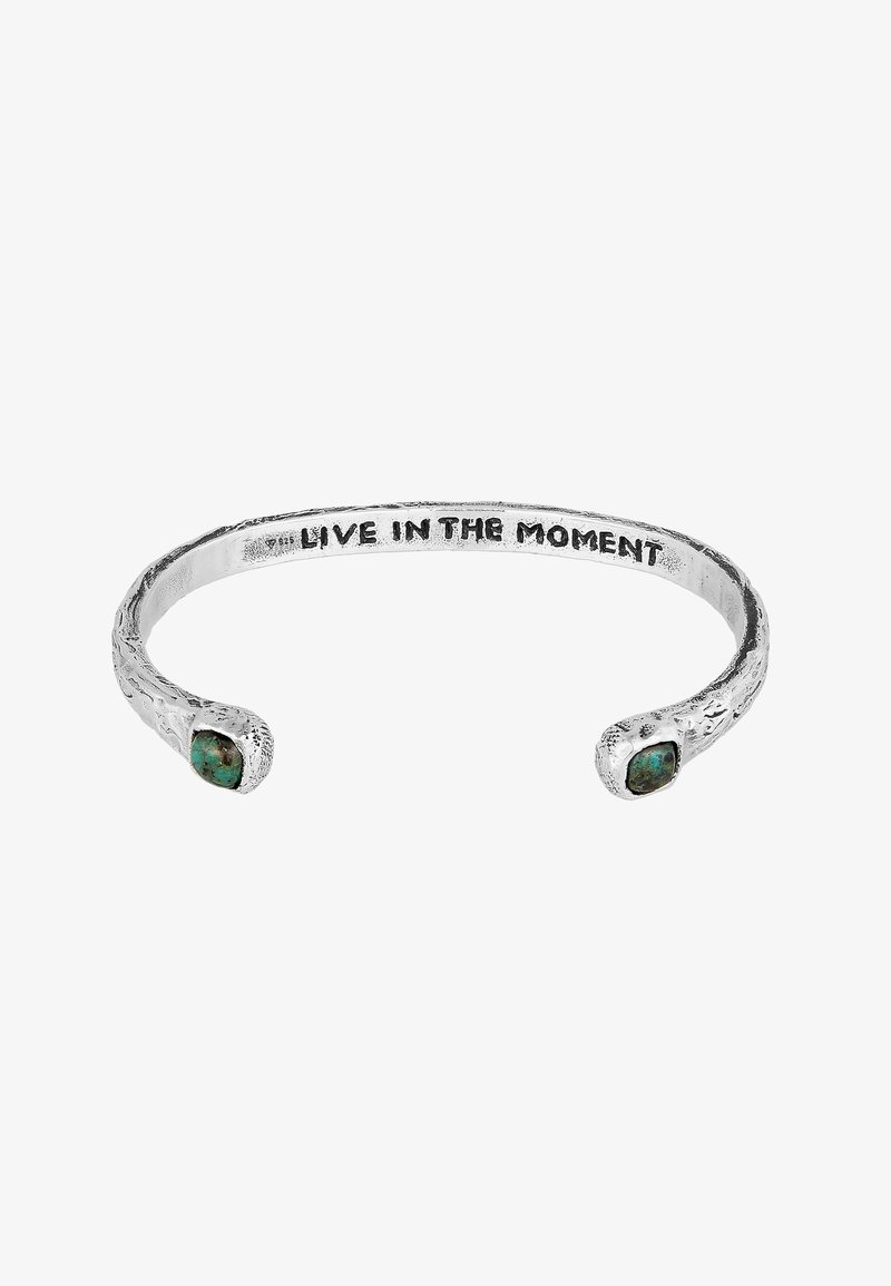 Sudraba aproce ar teksturētu virsmu, kuras katrā galā atrodas divi tīrkultūrs akmeņi un iegravēta frāze "Dzīvo mirklī".