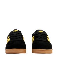 Zapatillas de ante negras con rayas amarillas, que cuentan con un diseño de cordones y suelas de goma. Textura suave y forma simple y redondeada.