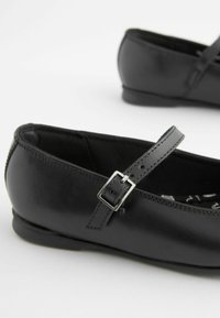 Svarta läderskor i ballerina-stil har en rund tå, en tunn rem med en fyrkantig silverspänne och en låg, platt klack för komfort.