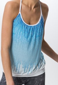 Kvinna i en blå och vit ärmlös halterneck-top med prickigt mönster och svarta och vita leggings, som står mot en enfärgad bakgrund.