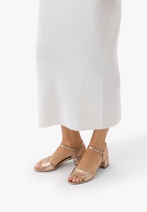 Stopy w sandałach na blokowym obcasie w kolorze metalicznego różowego złota z paskami wokół kostki, zestawione z białą, prążkowaną spódnicą midi.