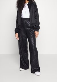 Svart bomberjacka med ribbade ärmar över vit skjorta, kombinerat med svarta mönstrade vida byxor och vita sneakers. Slät textur överlag.