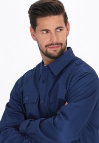 Cămașă bleumarin cu închidere butonată pe față, două buzunare pe piept și un guler clasic. Confecționată dintr-un material neted cu o croială ușor relaxată.
