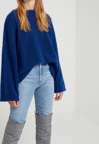 Blauer gestrickter Pullover mit weiten Ärmeln, kombiniert mit hellblauen Jeans und grauen karierten Stiefeln bis zum Knie. Weiche Textur mit lockerer Passform.