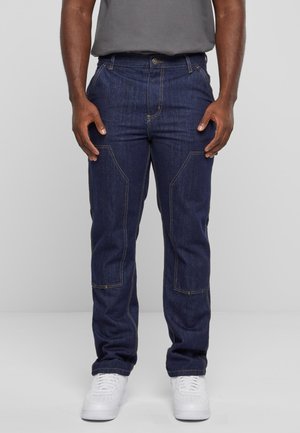 Uomo che indossa jeans di denim blu scuro con cuciture rinforzate e sneakers bianche, in piedi di fronte a uno sfondo grigio chiaro uniforme.