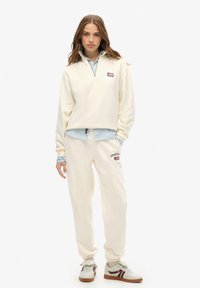Vrouw in een crèmekleurige sweatshirt en joggers met logo's, een lichtblauw shirt eronder, en witte sneakers met bruine en bordeaux details.