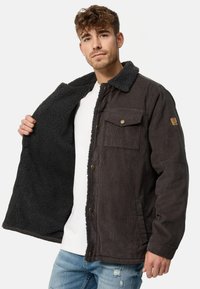 Donker corduroy jasje met een zachte, gewatteerde zwarte fleece voering, met knopen aan de voorkant en een borstzak. Draagt open over een wit t-shirt.